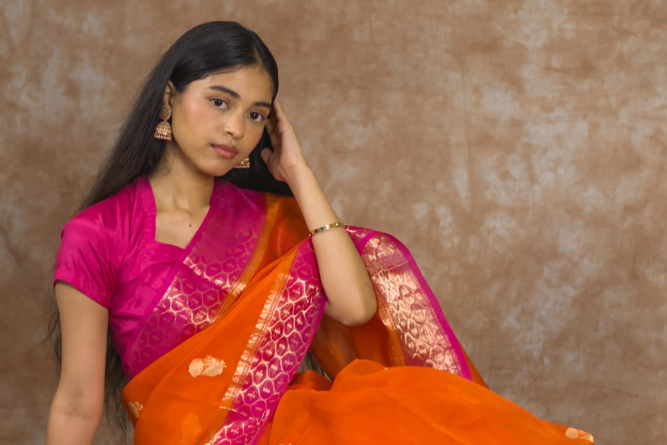 Woman wearing a pink blouse with an orange saree against a brown background